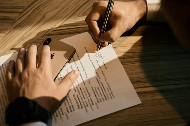 Eine Person schreibt auf einem Dokument, während eine andere Hand auf dem Tisch liegt.
