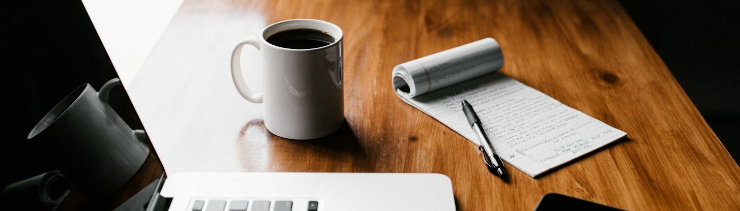 Ein Tisch mit einem Laptop, einer Tasse Kaffee, einem Notizblock und einem Stift.