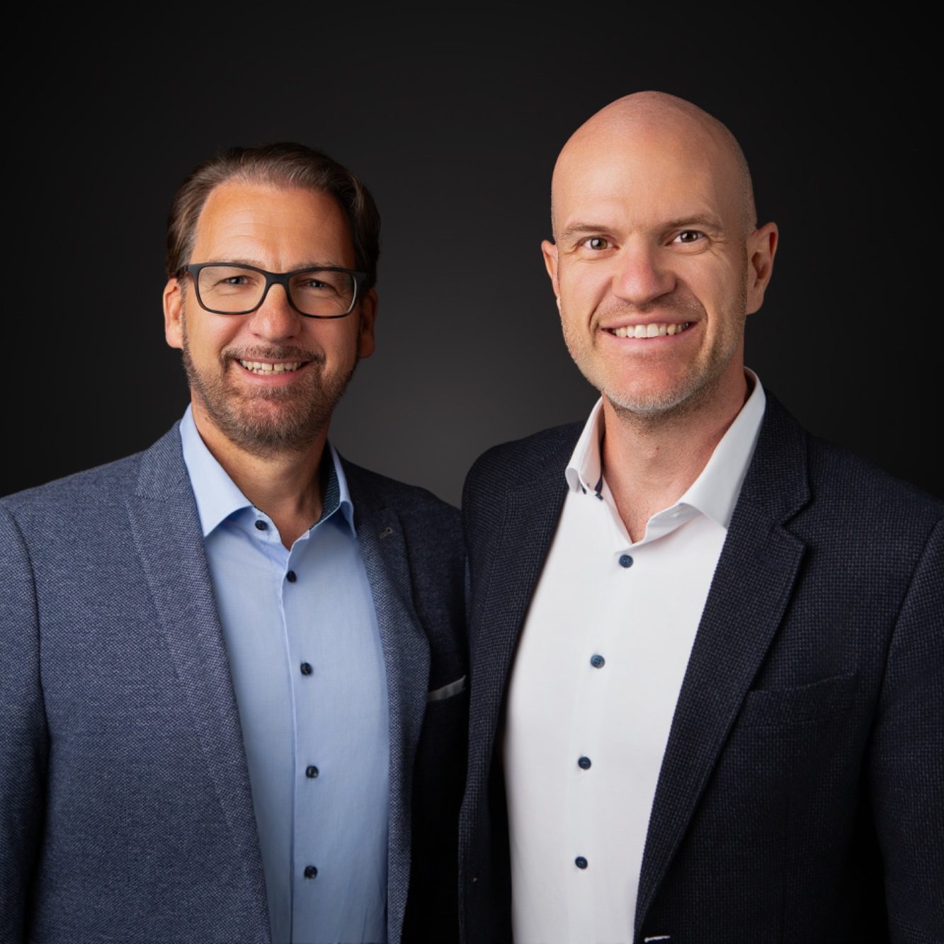 Stefan Bröcker und Oliver Greis Zwei Männer in Anzügen lächeln vor einem dunklen Hintergrund.